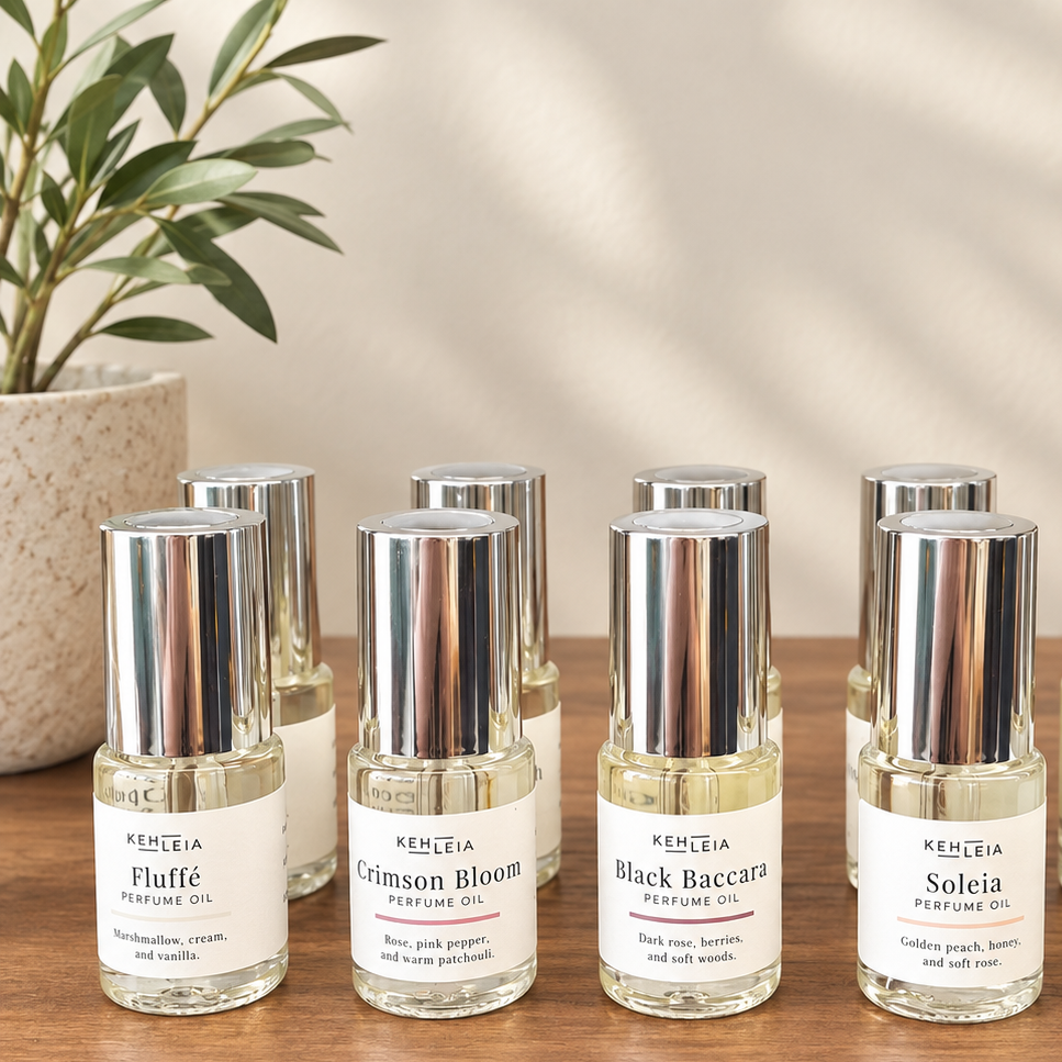Small perfume oil bottles with silver caps and white labels arranged on a wooden surface. The labels read KEHLEIA and the scents Fluffe, Crimson Bloom, Black Baccara, and Soleia.