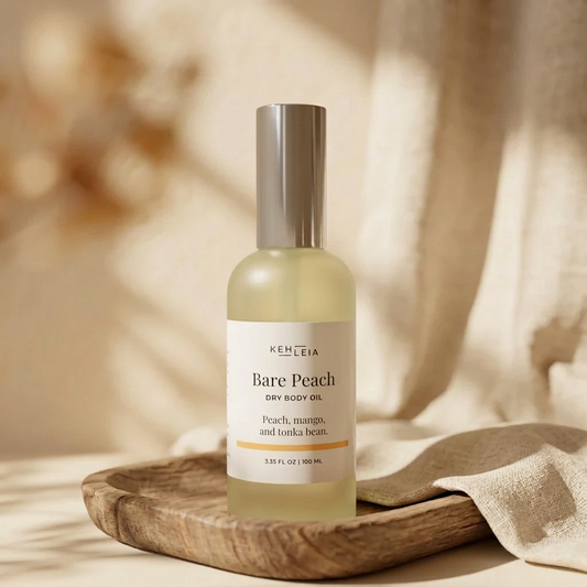 A bottle of Kehlea Bare Peach Dry Body Oil stands on a wooden tray with a soft beige fabric draped beside it. The bottle is frosted with pale yellow liquid and has a metallic silver cap, set against a warm, softly blurred background with light shadows.
