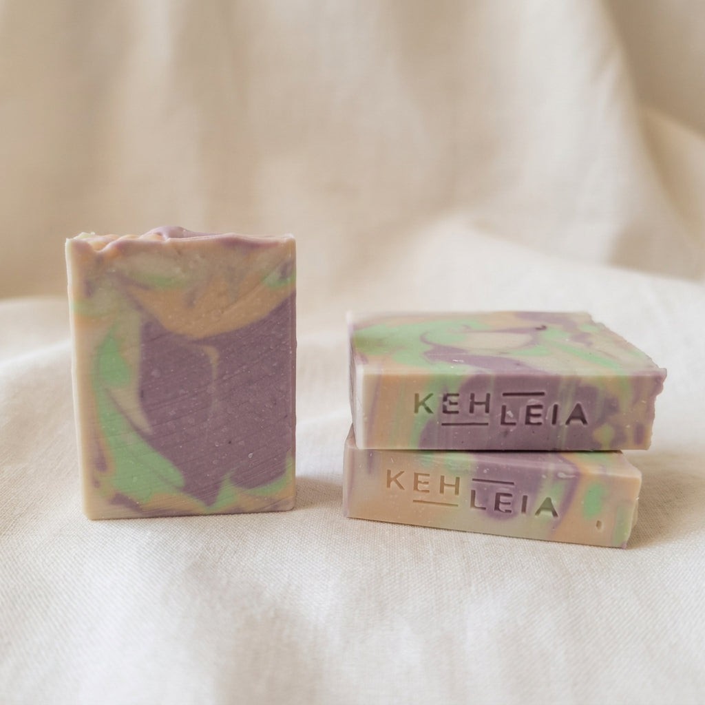 Three handmade marbled soaps in pastel purple, green, and beige tones, with two stacked soaps embossed with the brand name KEHLEIA.